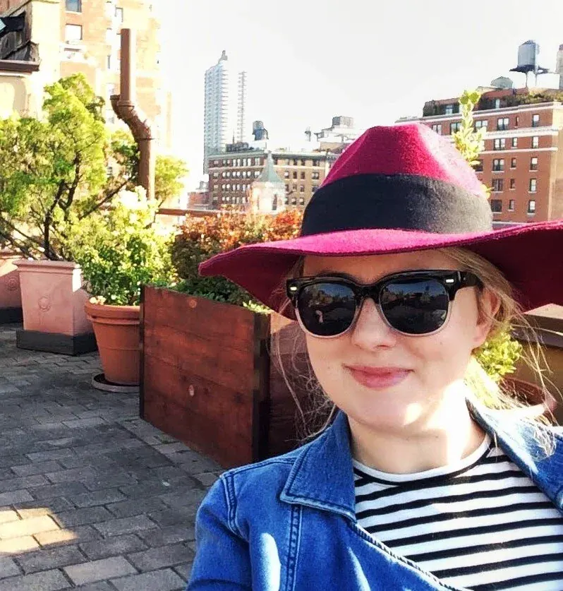 A woman wearing a pink and black wide-brimmed hat and black sunglasses, smiling outdoors on a rooftop terrace with potted plants, city buildings, and a partly cloudy sky in the background.