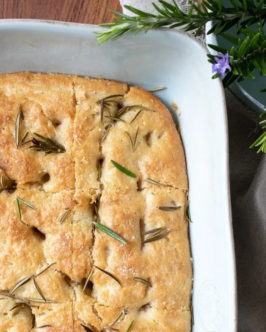 Golden sourdough focaccia topped with rosemary in a white baking dish